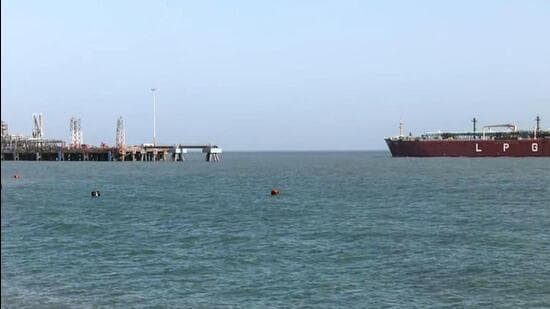 A large red LPG tanker vessel sailing across calm blue waters toward a port terminal with industrial docking structures in the background under a clear sky.